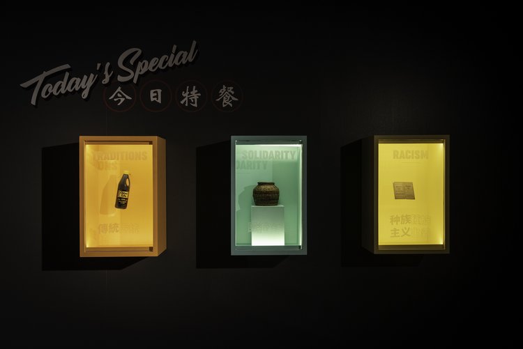 Black gallery wall with “Today’s Special / 今日特餐” lettered above three display cases holding contemporary and older condiment and food vessels and words, translucent, on the plexiglass front, including “Traditions / 傳統” (in Traditional Chinese); “Solidarity”; “Racism / 种族 主义” (in Simplified Chinese). Detail from A Seat at the Table exhibition at the Museum of Vancouver.