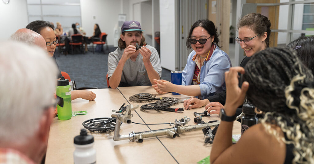 A group of people sitting around a desk looking at bicycle parts.