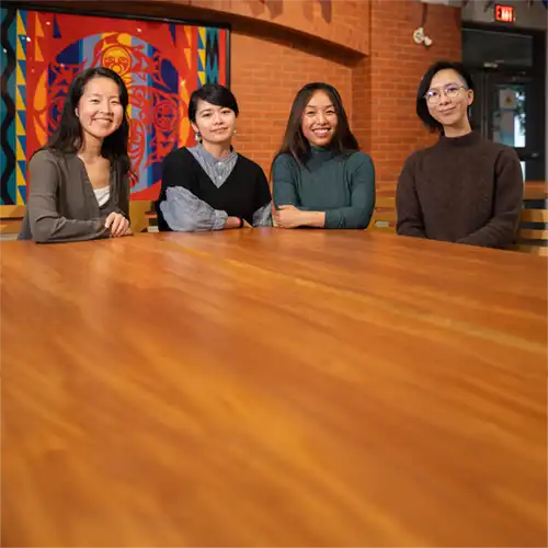 Four women of Asian descent sitting together at a wooden table looking at the camera.